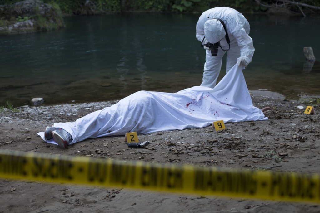 A photograph of a crime scene on a beach. An investigator is taking pictures of the body, wrapped in a white sheet. This was the scene at Gilgo Beach.