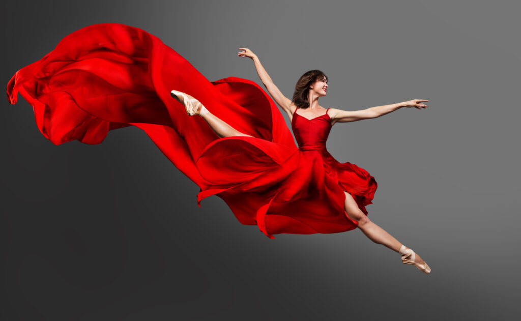 A female ballet dancer leaping as her billowing, bright red dress trails behind her.