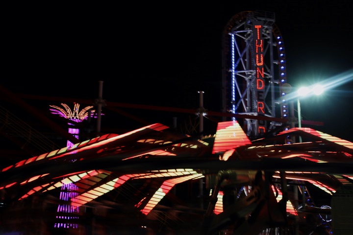 A spinning ride in the restored Luna Park lit up at night with the Thunderbolt and Parachute Jump lit in the background. 