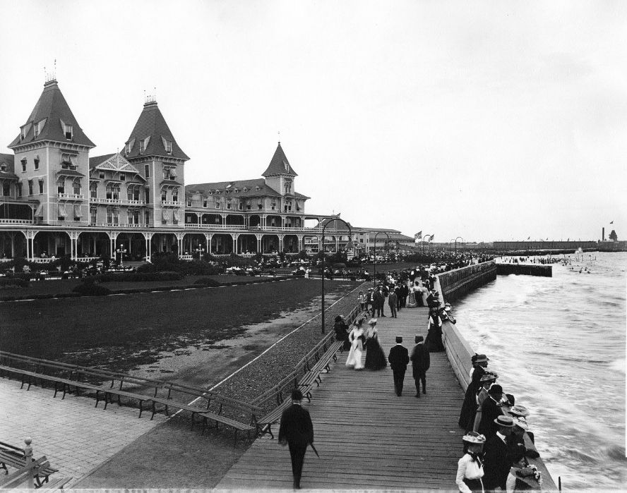 The Brighton Beach Hotel in the background with people strolling the boardwalk by the ocean. 